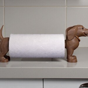 May include: A brown dachshund-shaped paper towel holder. The holder is made of a solid material and has a roll of white paper towels in the middle. The dog is standing on its four legs, with its tail up.