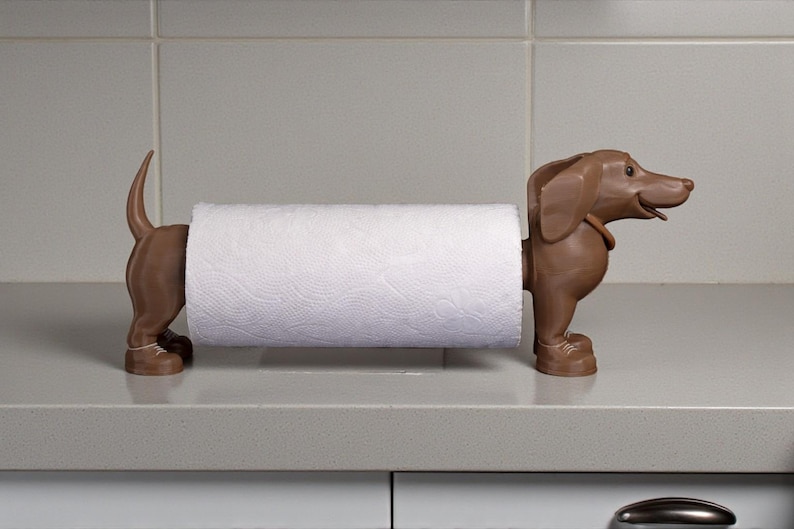 May include: A brown dachshund-shaped paper towel holder. The holder is made of a solid material and has a roll of white paper towels in the middle. The dog is standing on its four legs, with its tail up.