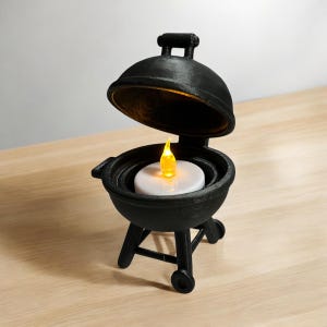 May include: A black miniature barbecue grill with the lid open, revealing a white candle with a flickering yellow flame. The grill has a handle, wheels, and a three-legged base. The candle is round and sits inside the grill.