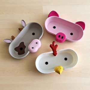 May include: Three animal-themed serving trays: a cow, a pig, and a chicken. The cow tray is gray with brown and pink accents. The pig tray is pink with dark pink ears and snout. The chicken tray is white with red and yellow accents.