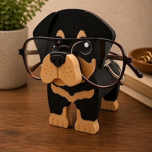 May include: A wooden dog-shaped eyeglass holder in black and tan. The holder is designed to hold eyeglasses, with the glasses resting on the dog's nose. The dog has a friendly expression and is wearing a pair of brown-rimmed glasses.