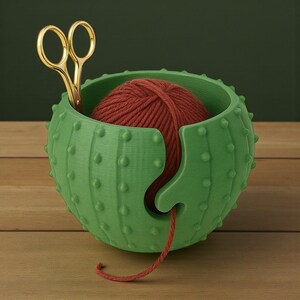 May include: A green cactus-shaped yarn bowl with a red yarn ball inside. Gold scissors are placed in the bowl. The bowl has a slot to feed the yarn through. The bowl is on a wooden surface.