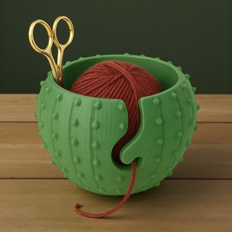 May include: A green cactus-shaped yarn bowl with a red yarn ball inside. Gold scissors are placed in the bowl. The bowl has a slot to feed the yarn through. The bowl is on a wooden surface.