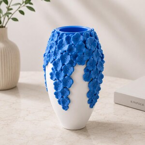 May include: A white ceramic vase with a blue interior and a cascade of blue floral embellishments. The vase has a smooth, glossy finish and a unique, textured design. The vase is a decorative home decor item.