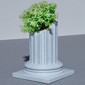 May include: A light grey, fluted column planter with a square base, holding a vibrant green plant. The planter has a classic architectural design. The plant's bright green leaves contrast with the column's neutral colour. A decorative item for the home.