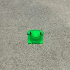 May include: A bright green frog figurine with black eyes. The frog is sitting on a grey surface.