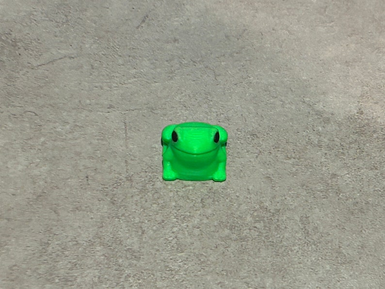 May include: A bright green frog figurine with black eyes. The frog is sitting on a grey surface.