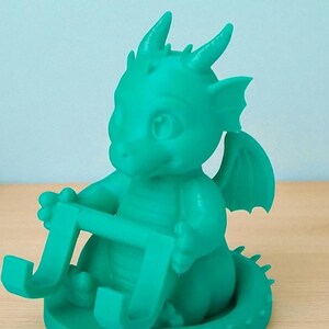 May include: A teal-colored dragon figurine holding a phone or tablet stand. The dragon has large eyes, small horns, and wings. The stand is a curved shape, designed to hold a device. The figurine is made of a smooth material and sits on a round base.
