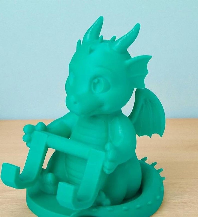 May include: A teal-colored dragon figurine holding a phone or tablet stand. The dragon has large eyes, small horns, and wings. The stand is a curved shape, designed to hold a device. The figurine is made of a smooth material and sits on a round base.
