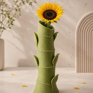 Puede incluir: Un jarr&oacute;n verde con forma de bamb&uacute; sostiene un girasol amarillo brillante. El jarr&oacute;n tiene una superficie texturizada y detalles de hojas. P&eacute;talos amarillos dispersos completan la decoraci&oacute;n natural.