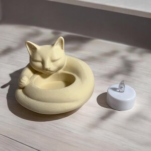 May include: A cream-colored cat-shaped candle holder with a sleeping cat design. The cat curls around a circular opening for a candle. A small, white, battery-operated tea light sits next to the holder.