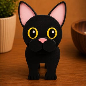 May include: A black cat figurine with large yellow eyes, pink inner ears, and a pink nose. The cat is standing upright with its front paws visible. The figurine is made of a matte black material and is set against a warm brown background.