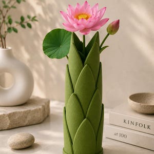 May include: A green felt vase with a lotus flower and bud. The pink lotus flower has a yellow center and green leaf. The vase is cylindrical with a layered petal design. A white vase and a stone are in the background.