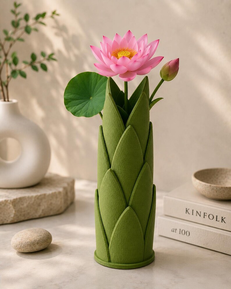 May include: A green felt vase with a lotus flower and bud. The pink lotus flower has a yellow center and green leaf. The vase is cylindrical with a layered petal design. A white vase and a stone are in the background.