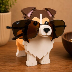 May include: A dog-shaped eyeglass holder in brown, white, and orange. The dog is wearing black sunglasses with the Ray-Ban logo. The holder is made of a sturdy material and is designed to hold eyeglasses.