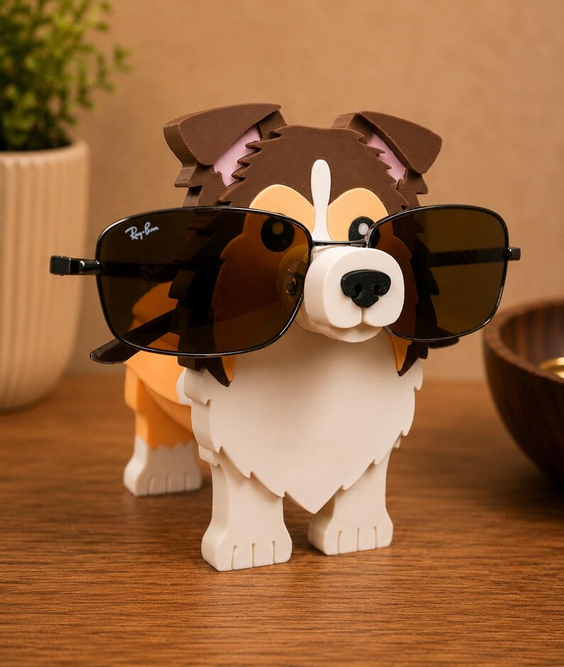 May include: A dog-shaped eyeglass holder in brown, white, and orange. The dog is wearing black sunglasses with the Ray-Ban logo. The holder is made of a sturdy material and is designed to hold eyeglasses.