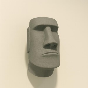 May include: A gray, textured Moai statue head, a replica of the iconic Easter Island statues. The sculpture features a large nose, prominent chin, and detailed facial features. The statue is mounted on a light-colored wall.