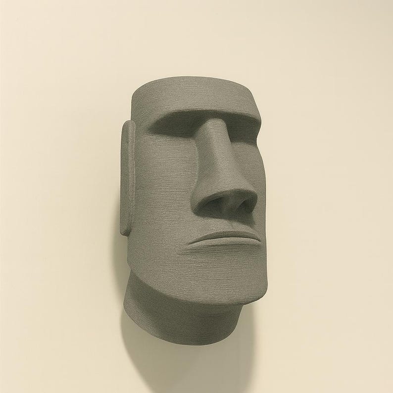 May include: A gray, textured Moai statue head, a replica of the iconic Easter Island statues. The sculpture features a large nose, prominent chin, and detailed facial features. The statue is mounted on a light-colored wall.