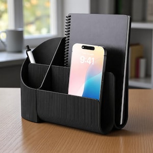 May include: Black desk organizer with multiple compartments. It holds a spiral notebook, a smartphone displaying 9:41, and a pen. The organizer has a ribbed texture and sits on a wooden surface.