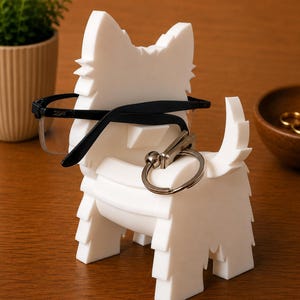 May include: A white, dog-shaped eyeglasses holder with black-framed glasses resting on it. A silver keychain hangs from the holder. The holder is on a wooden surface, with a small plant and a wooden bowl in the background.