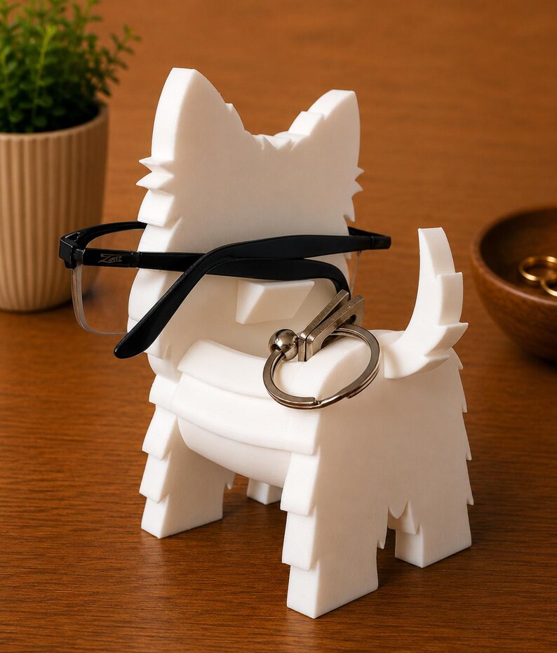 May include: A white, dog-shaped eyeglasses holder with black-framed glasses resting on it. A silver keychain hangs from the holder. The holder is on a wooden surface, with a small plant and a wooden bowl in the background.