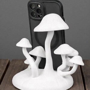 May include: A white mushroom-shaped phone stand with multiple mushroom caps and stems. The stand is designed to hold a black smartphone. The stand is made of a textured material and sits on a wooden surface.