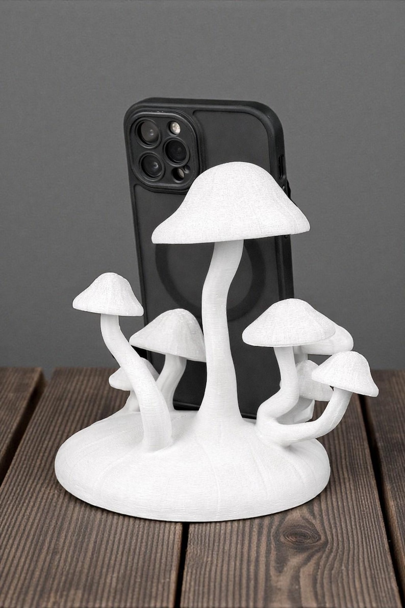 May include: A white mushroom-shaped phone stand with multiple mushroom caps and stems. The stand is designed to hold a black smartphone. The stand is made of a textured material and sits on a wooden surface.