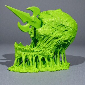 May include: A neon green, 3D-printed sculpture of a grotesque, open-mouthed creature with a spear piercing its head. The sculpture has a textured, organic appearance with dripping, viscous elements, set against a gray background.