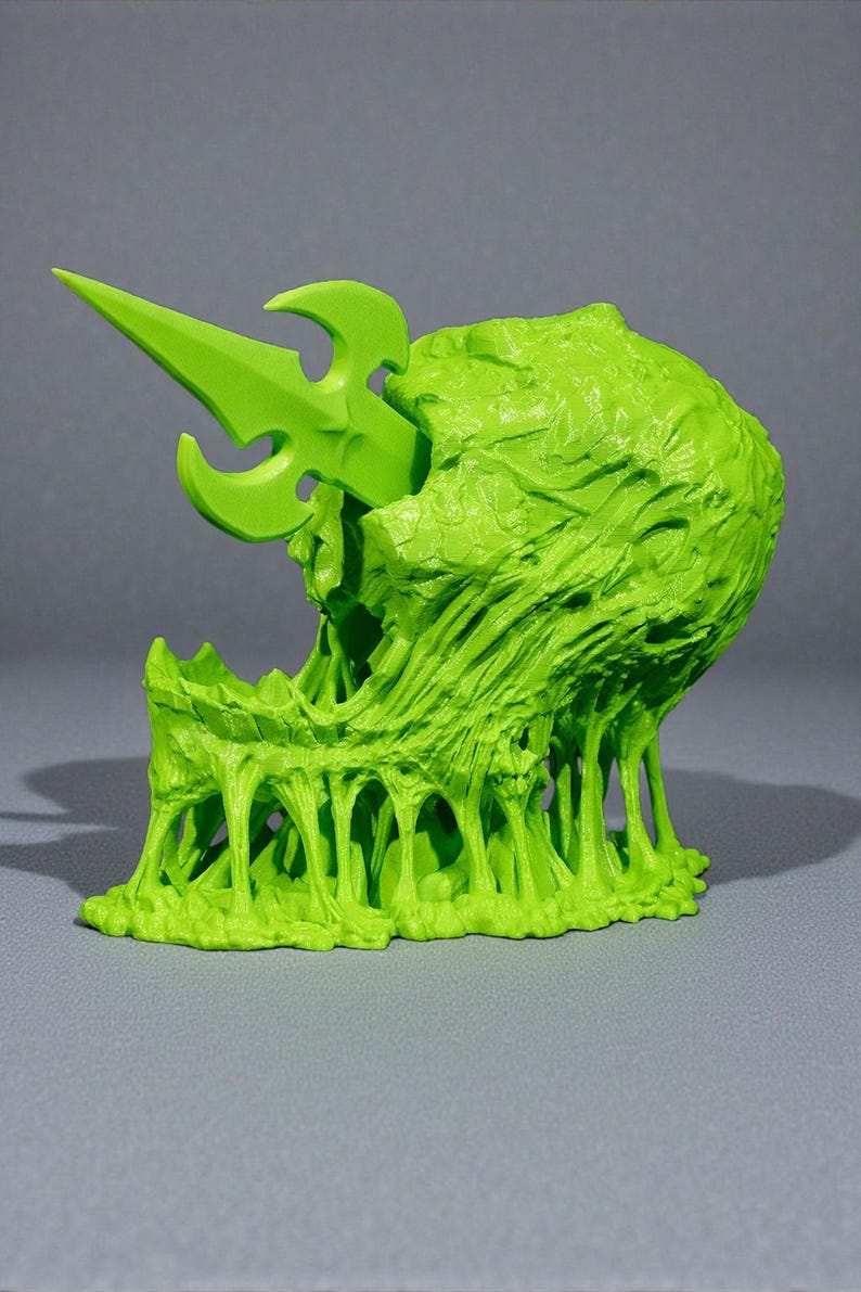 May include: A neon green, 3D-printed sculpture of a grotesque, open-mouthed creature with a spear piercing its head. The sculpture has a textured, organic appearance with dripping, viscous elements, set against a gray background.