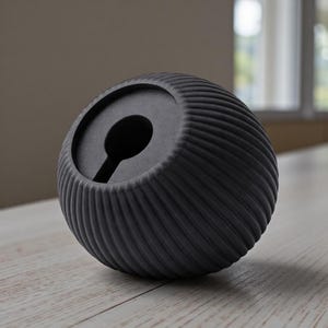 May include: A dark gray, spherical bottle stopper with a textured, ridged surface. The stopper has a keyhole-shaped opening on top. The object is sitting on a light-colored wooden surface.