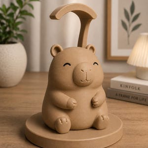 May include: A tan-coloured bear-shaped desk lamp. The lamp has a curved handle and sits on a round base. The bear has a smiling face with small, black eyes. The lamp is made of a textured material and is designed for a desk or bedside table.