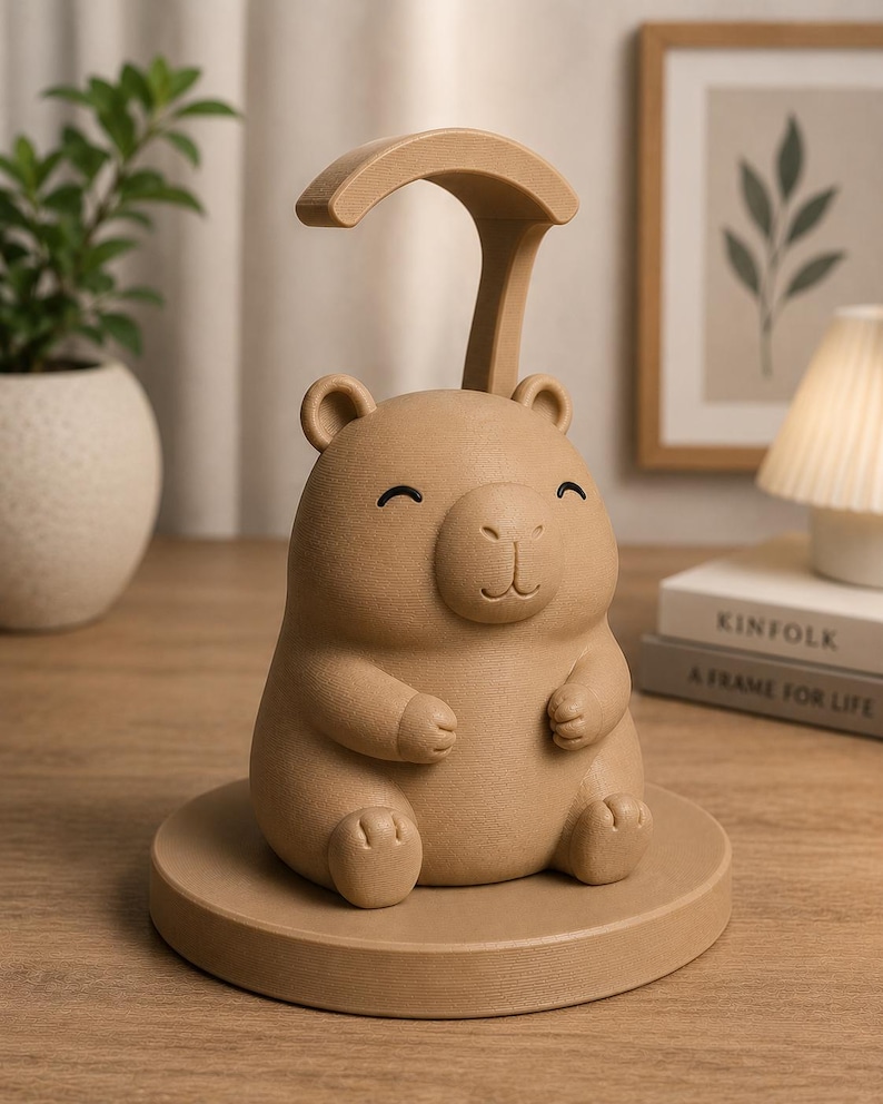 May include: A tan-coloured bear-shaped desk lamp. The lamp has a curved handle and sits on a round base. The bear has a smiling face with small, black eyes. The lamp is made of a textured material and is designed for a desk or bedside table.
