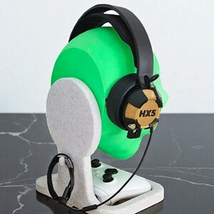 May include: A gaming headset with the text "HX5" rests on a green head-shaped stand. The stand is white and holds a white game controller. The headset is black and gold.