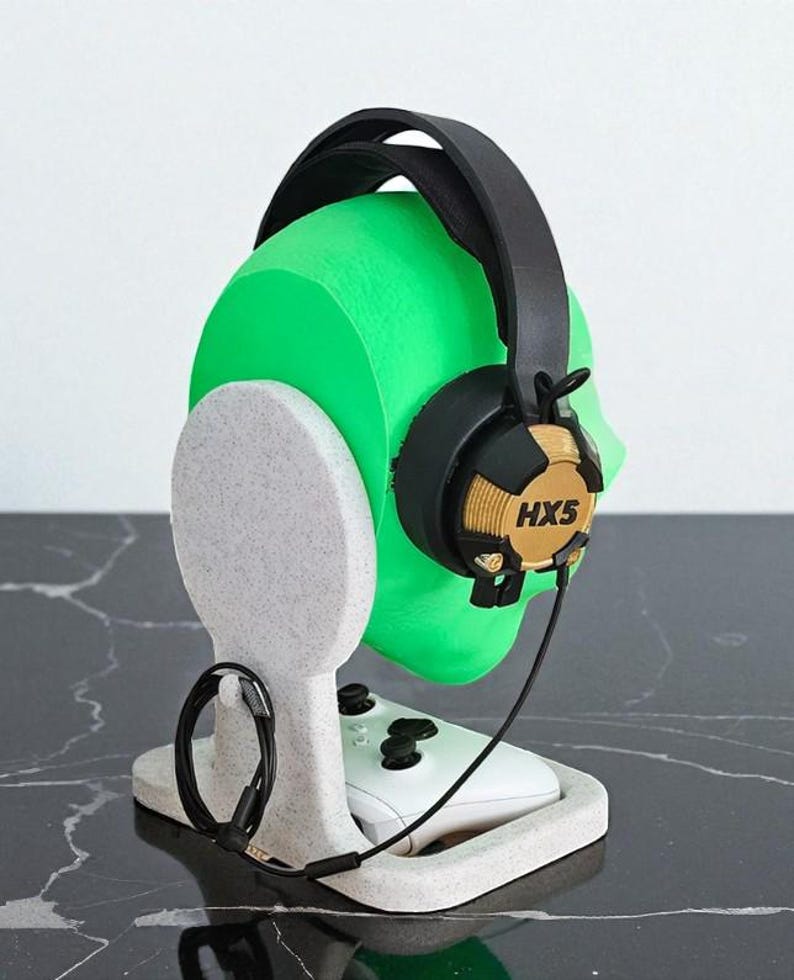 May include: A gaming headset with the text "HX5" rests on a green head-shaped stand. The stand is white and holds a white game controller. The headset is black and gold.
