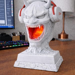May include: A white, detailed sculpture of a demonic head with horns and an open mouth, illuminated with an orange glow. The sculpture wears white and red headphones. The base is square and textured.