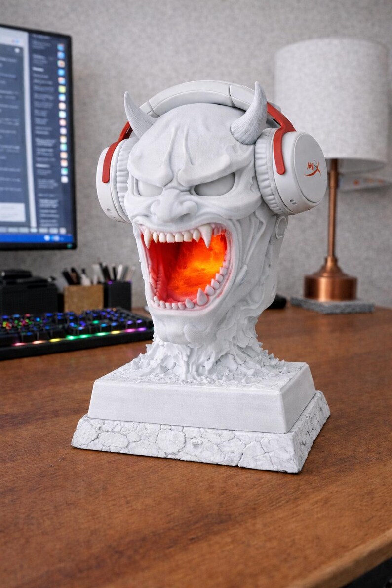 May include: A white, detailed sculpture of a demonic head with horns and an open mouth, illuminated with an orange glow. The sculpture wears white and red headphones. The base is square and textured.