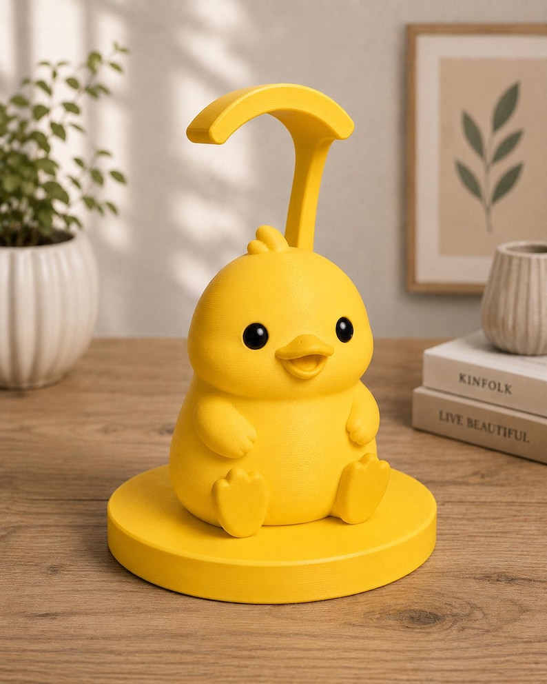May include: A yellow chick-shaped desk lamp with a curved top and a round base. The lamp has black eyes and a smiling beak. The lamp is sitting on a wooden surface. The lamp is a decorative item.