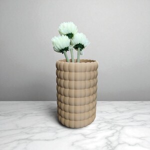 Pu&ograve; includere: Un vaso beige strutturato con un design arrotondato e impilato, che contiene tre fiori bianchi con steli verdi. Il vaso &egrave; appoggiato su una superficie di marmo, con uno sfondo neutro.