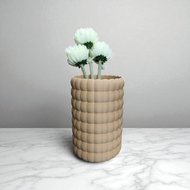 Pu&ograve; includere: Un vaso beige strutturato con un design arrotondato e impilato, che contiene tre fiori bianchi con steli verdi. Il vaso &egrave; appoggiato su una superficie di marmo, con uno sfondo neutro.