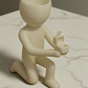 May include: A cream-colored figurine of a person kneeling, holding a small open box with a butterfly on top. The figurine has a rounded head with a hollowed-out top, and is on a white marble surface.