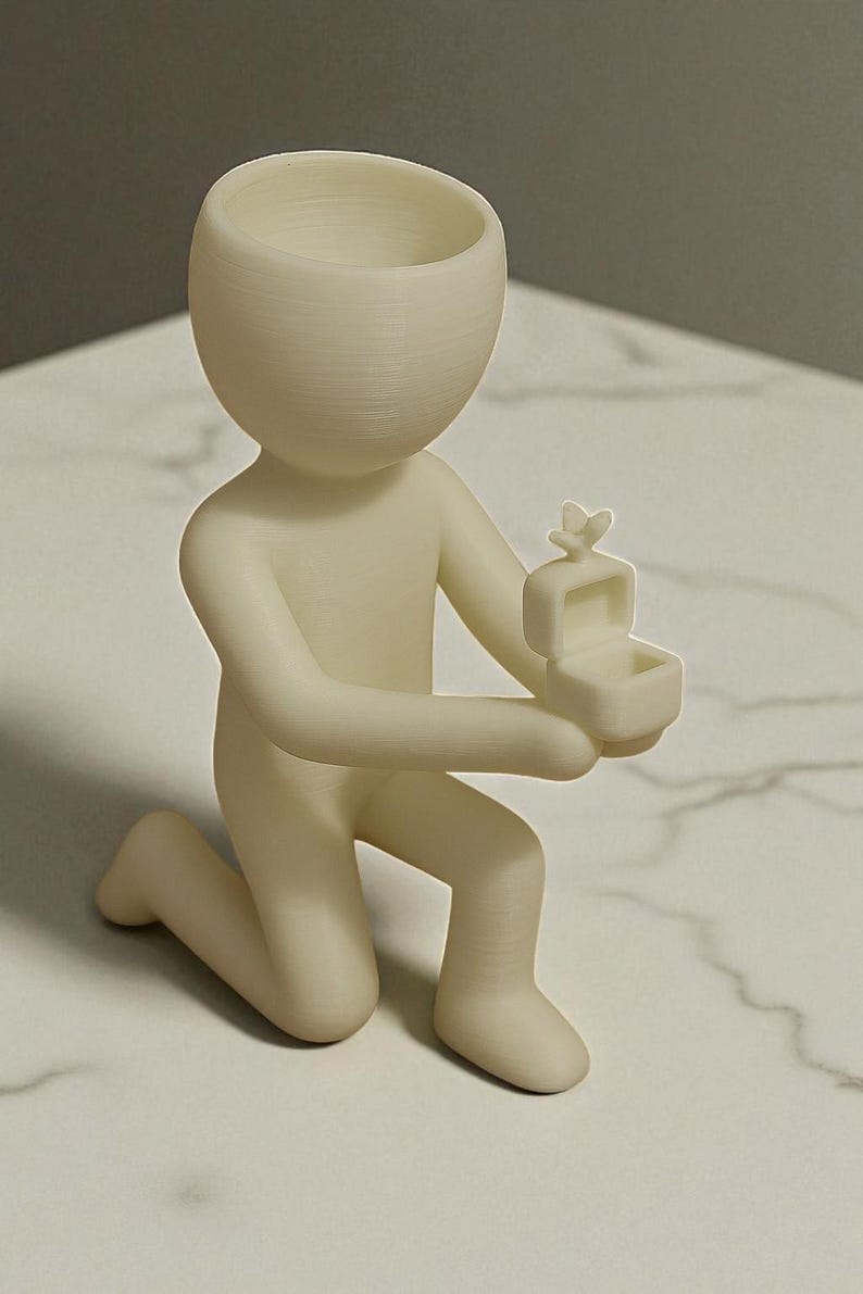 May include: A cream-colored figurine of a person kneeling, holding a small open box with a butterfly on top. The figurine has a rounded head with a hollowed-out top, and is on a white marble surface.