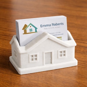 May include: A white house-shaped business card holder with a stack of business cards inside. The business cards have the name "Emma Roberts" and the title "Real Estate Agent" printed on them. The holder has a door and windows.
