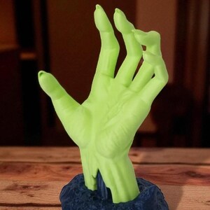 Puede incluir: Una escultura de mano verde con una superficie texturizada, que emerge de una base azul oscuro. La mano est&aacute; en un gesto, con los dedos ligeramente curvados. La escultura es probablemente un objeto decorativo o una obra de arte &uacute;nica.