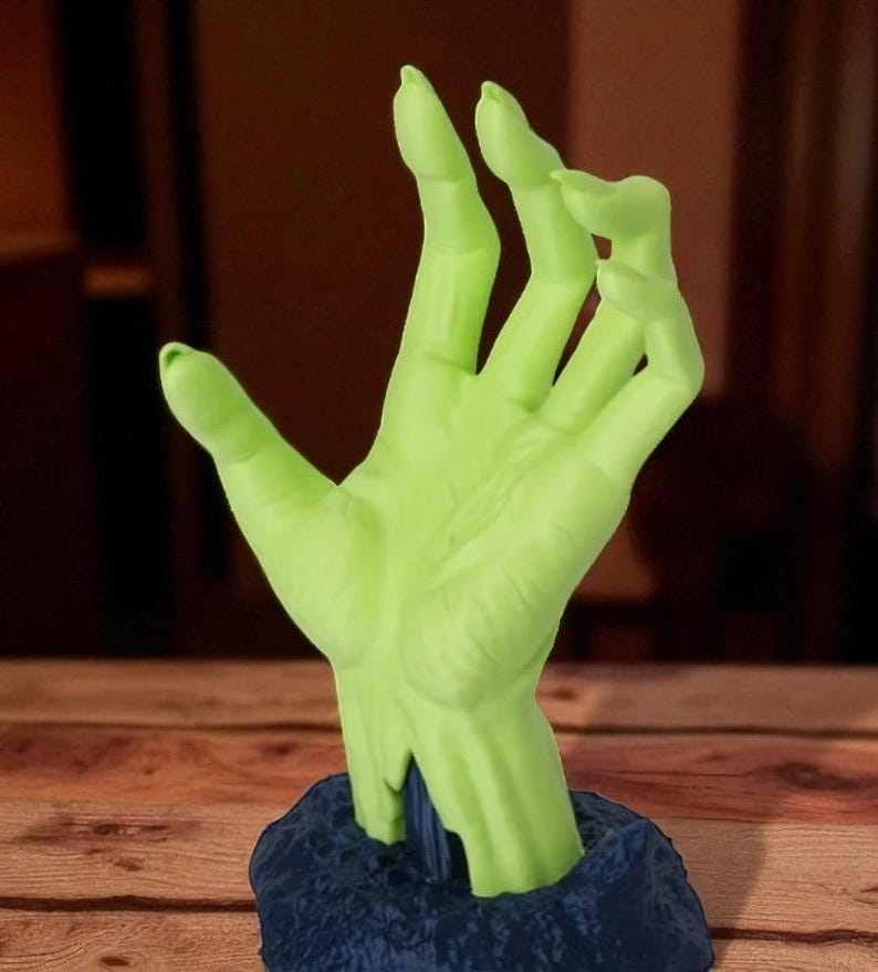 Puede incluir: Una escultura de mano verde con una superficie texturizada, que emerge de una base azul oscuro. La mano est&aacute; en un gesto, con los dedos ligeramente curvados. La escultura es probablemente un objeto decorativo o una obra de arte &uacute;nica.