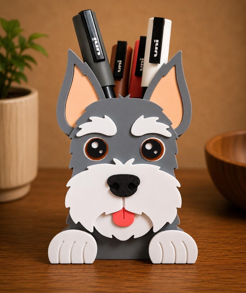 May include: A gray and white dog-shaped desk organizer holding several black, brown, and white pens. The organizer has a cute design with large eyes, a black nose, and a red tongue. The ears are a light orange color. The organizer is sitting on a wooden surface.