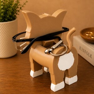 May include: A tan cat-shaped eyeglasses and key holder with black eyeglasses and a silver key ring. The cat has white accents on its paws and a white spot on its rear. A small plant and a book are in the background.