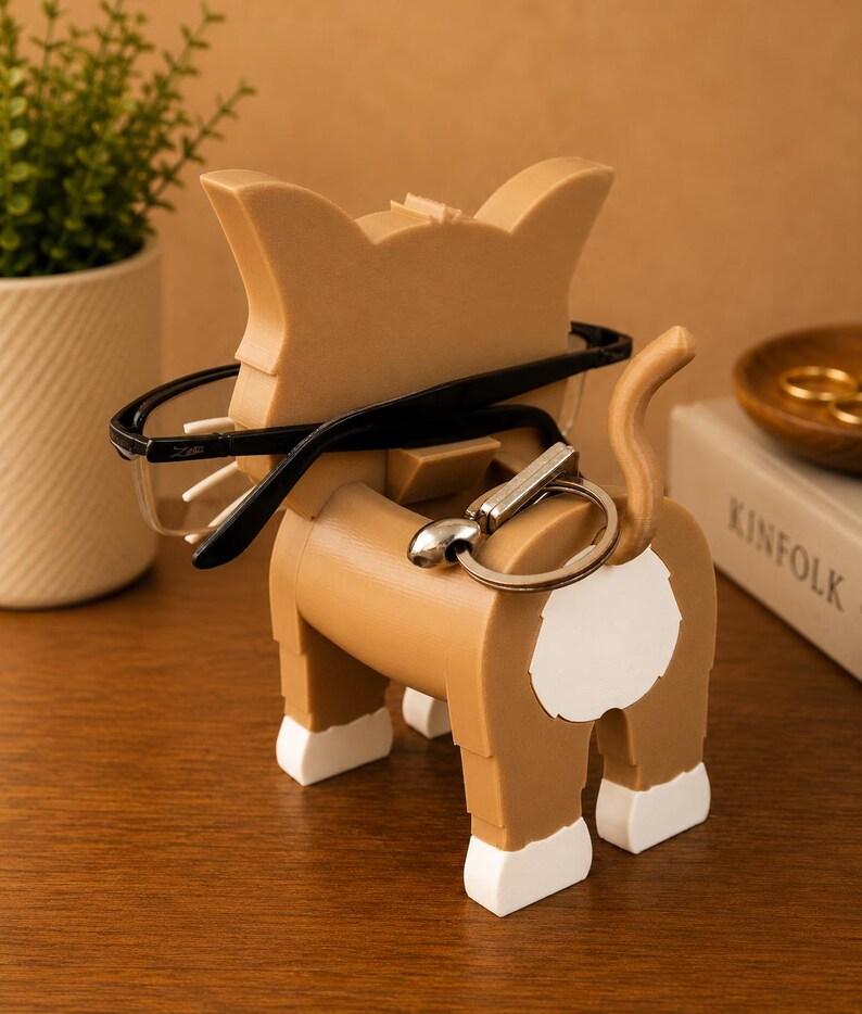 May include: A tan cat-shaped eyeglasses and key holder with black eyeglasses and a silver key ring. The cat has white accents on its paws and a white spot on its rear. A small plant and a book are in the background.