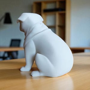 May include: A white ceramic bulldog figurine sitting on a wooden table. The dog is in a seated position, with its head turned to the side. The figurine has smooth, rounded features and is a solid, matte white color. The background is blurred, showing a room with tables and a shelf.