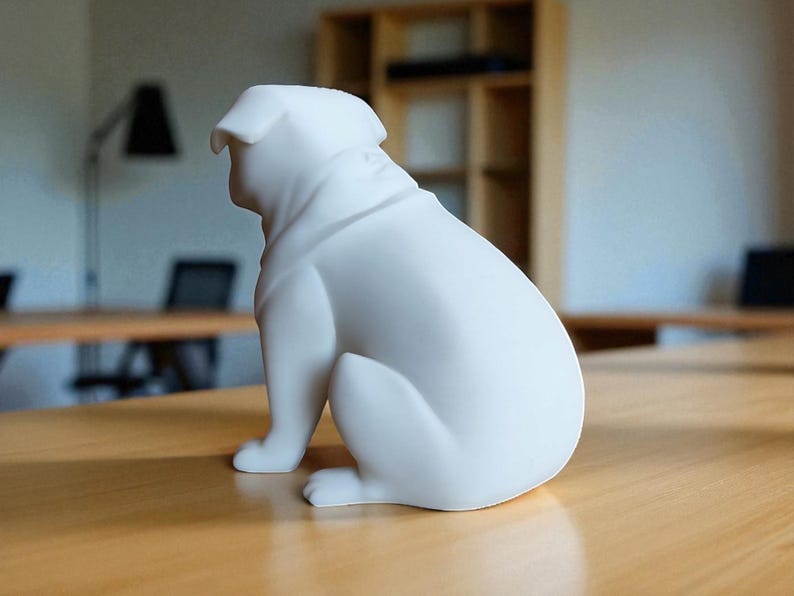 May include: A white ceramic bulldog figurine sitting on a wooden table. The dog is in a seated position, with its head turned to the side. The figurine has smooth, rounded features and is a solid, matte white color. The background is blurred, showing a room with tables and a shelf.