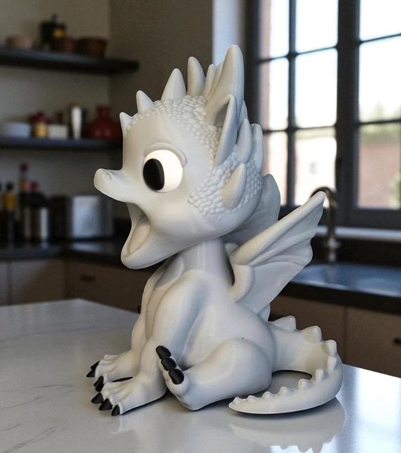 May include: A white dragon figurine with large eyes and an open mouth. The dragon has detailed scales, wings, and black claws. It is sitting on a white surface, with a kitchen in the background.
